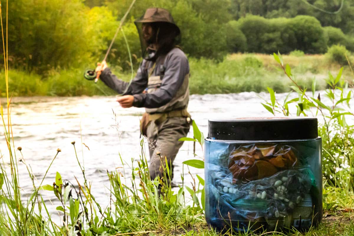 A man fly fishing in the background with a bear canister in the foreground