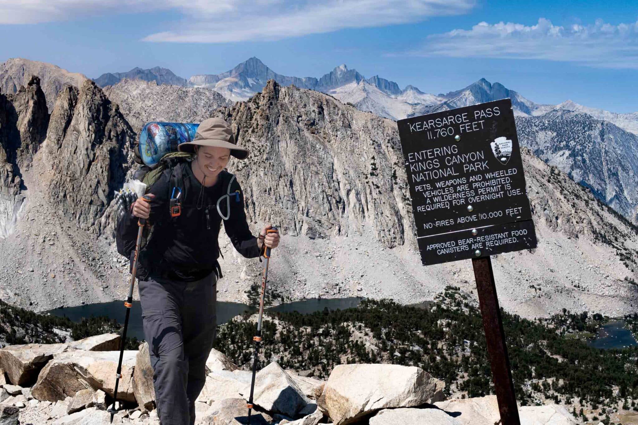 A man hikes over a mountain pass with a bear canister on his back