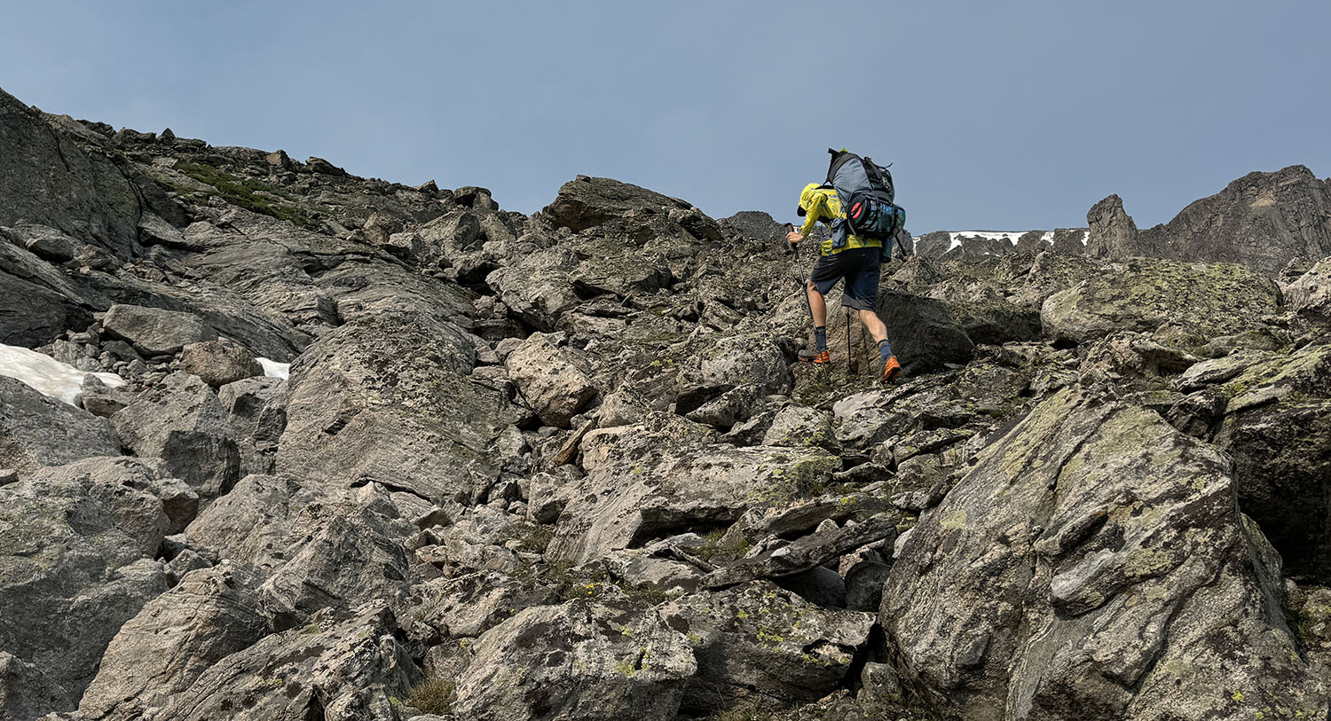 A hiker climbs up a steep and rocky slope with a bear proof canister strapped to the bottom of the pack