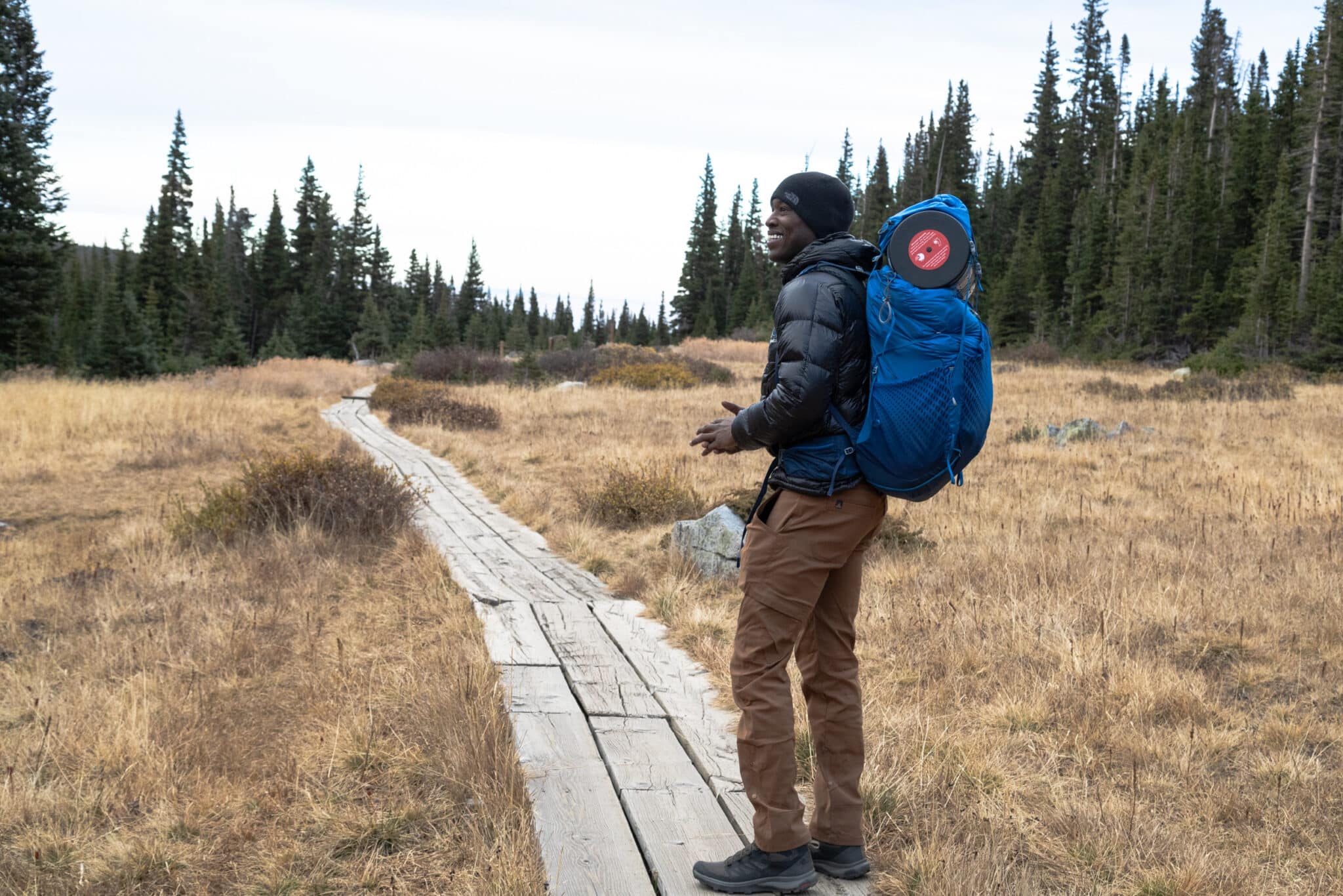 A man wearing a backpacking backapck smiles while standing on a bridge through a meadow